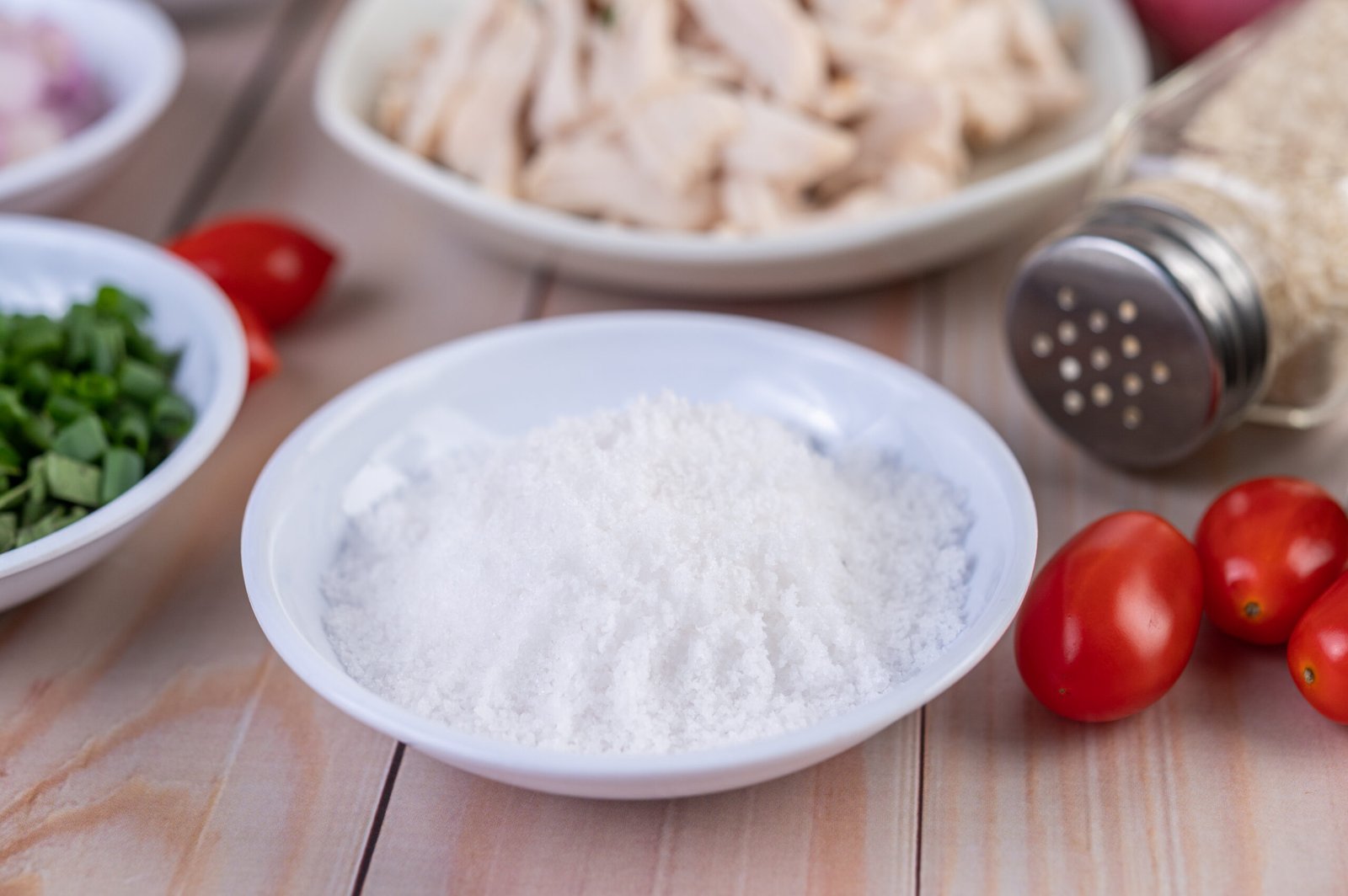 Salt in a white spoon, tomatoes placed on a wooden table. Selective focus.